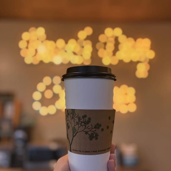 Hand holding a white coffee cup with a tree-patterned sleeve against blurred golden lights.