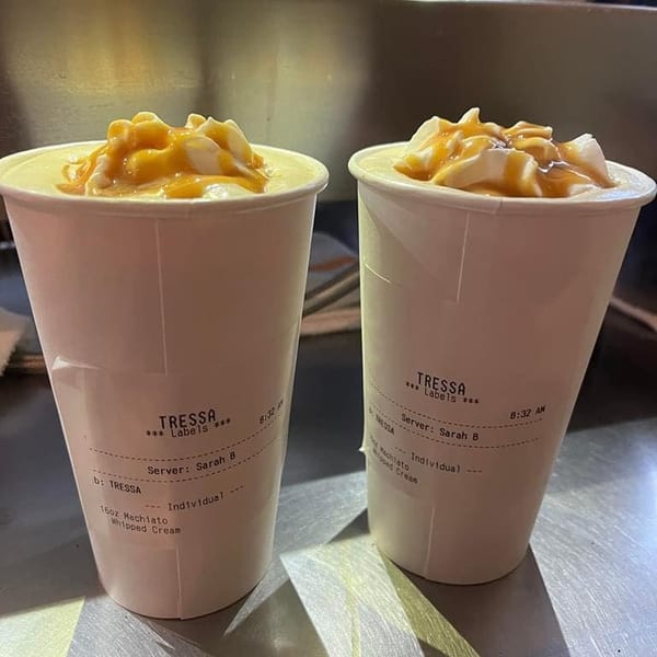 Two Tressa Labels macchiatos with whipped cream and caramel drizzle on a metal counter.