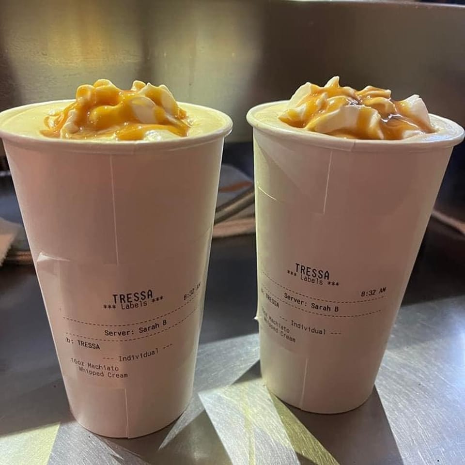 Two Tressa Labels macchiatos with whipped cream and caramel drizzle on a metal counter.