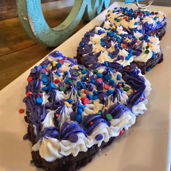 Three heart-shaped brownies with purple and white frosting and colorful sprinkles on a white platter.