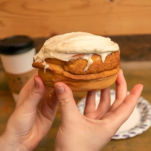 Hands holding a large, frosted cinnamon roll with a coffee cup and paper plate in the background.