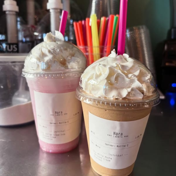 Two Nora brand blended drinks with whipped cream and pink straws on a counter.