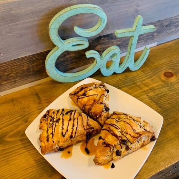 Three chocolate chip scones drizzled with caramel and chocolate on a white plate next to a blue "Eat" sign.