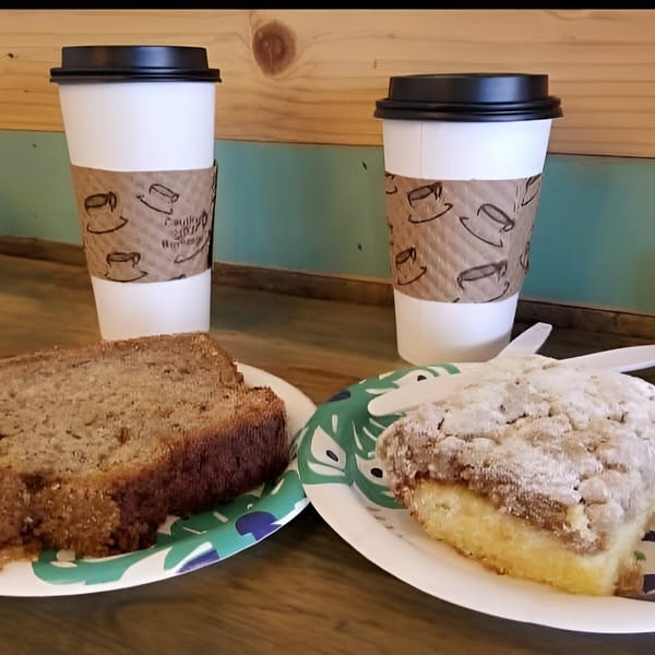 Two coffee cups with banana bread and coffee cake on paper plates on a wooden table.