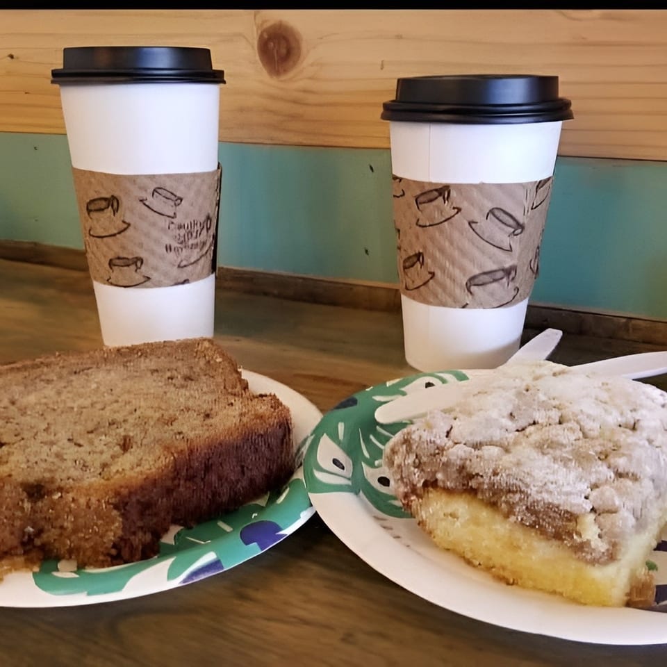 Two coffee cups with banana bread and coffee cake on paper plates on a wooden table.