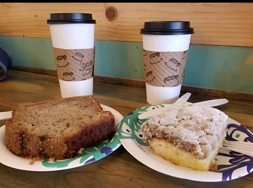 Two coffee cups with banana bread and coffee cake on paper plates on a wooden table.