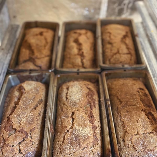 Six freshly baked loaves of banana bread in metal pans, topped with sparkling sugar.
