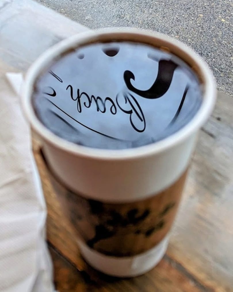 A cup of coffee with "Beach" reflected on its surface, sitting on a wooden table.