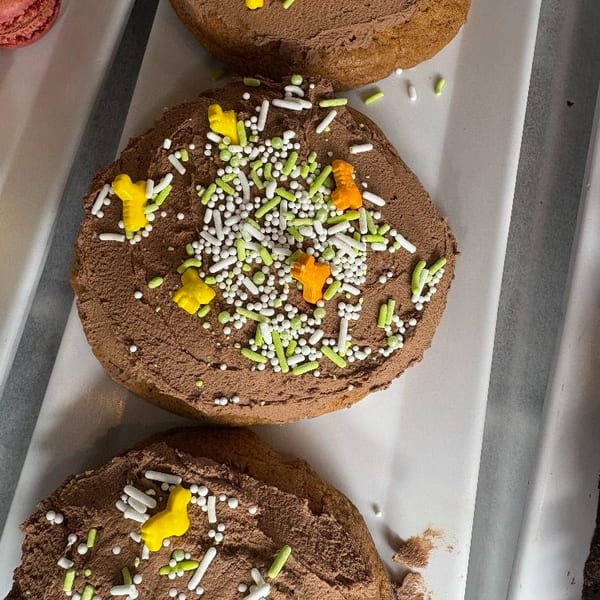 Three chocolate frosted cookies with green, white, yellow, and orange sprinkles on a white tray.
